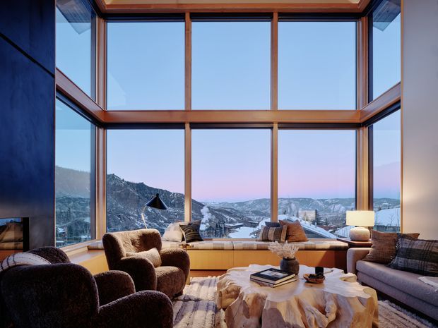 A sophisticated living room with expansive double-height timber-framed glazing overlooking a snowy mountainous landscape. The room features a dark stone or plaster fireplace wall, warm wood window framing and built-in window seating, plush brown lounge chairs, a grey sofa, layered textiles, and a sculptural natural timber coffee table. The design combines a luxurious lodge-like atmosphere with strong contemporary architectural glazing and a dramatic alpine outlook.