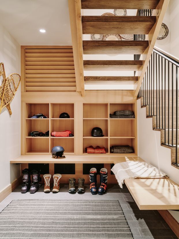 A bright, refined ski-entry mudroom beneath an open-riser timber staircase. The space features extensive light timber built-in shelving and cabinetry, a long bench with concealed storage, black metal stair balustrading, ski and snowboard gear, boots, helmets, woven wall decor, and a striped floor rug. Sunlight enters from the right, creating a warm and functional alpine-oriented entry area.
