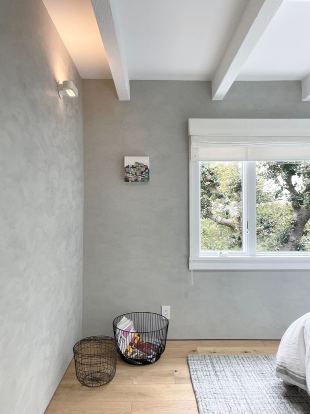 A bright, compact bedroom corner with pale grey textured walls, white exposed ceiling beams, a timber floor, and a large white-framed window with a Roman blind. The scene includes the edge of a bed, a grey rug, wire baskets, a wall light, and a small artwork, creating a simple contemporary interior.