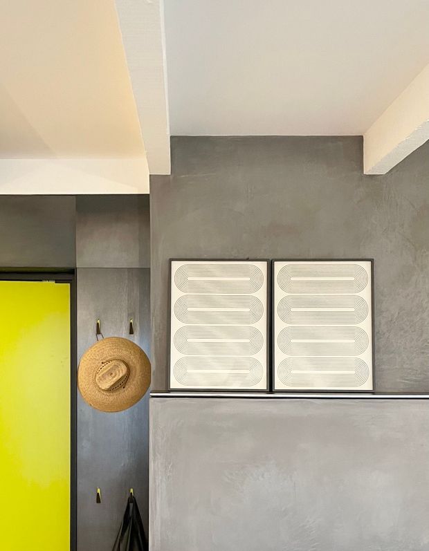 A narrow contemporary interior passage or entry area with smooth grey plaster or concrete-look walls, white ceiling beams, and a vivid yellow door or panel at left. Two matching framed abstract line artworks sit on a slim ledge above a grey half-height wall. A woven straw hat hangs beside the doorway, with dark hanging garments below. The space has a restrained industrial-modern palette with a bright yellow accent.