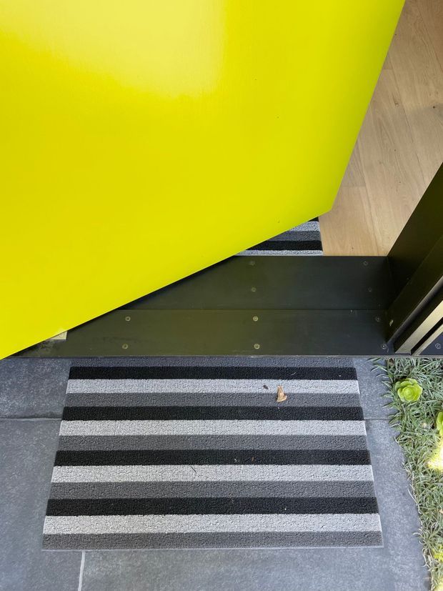 Close-up view of a bold residential entryway, showing a bright yellow door, dark metal threshold and framing, striped black-and-grey doormat, dark paving, and a small planted edge. The image focuses on the entrance flooring and door details.