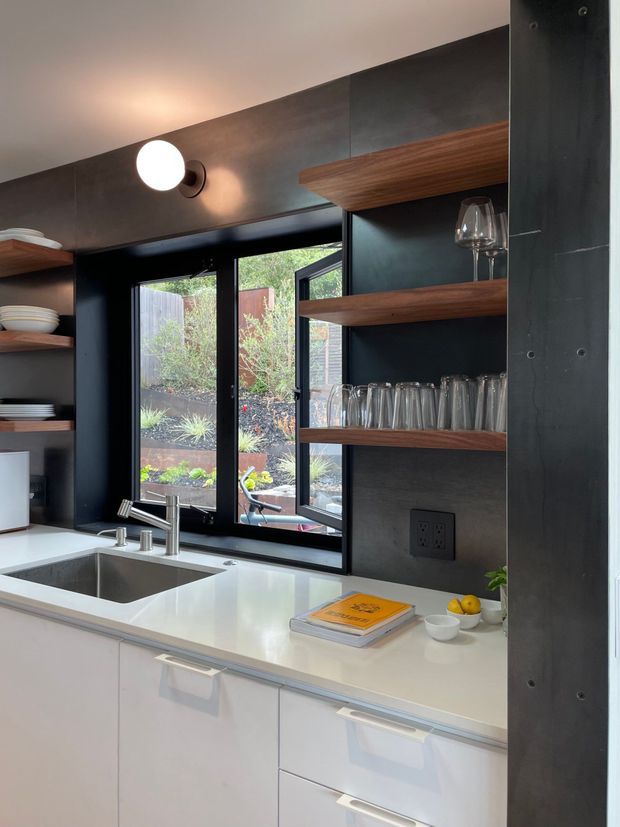 A compact contemporary kitchen with a white engineered-looking benchtop, integrated sink and chrome tapware, glossy white lower cabinetry, and dark charcoal wall panels and shelving. Warm timber open shelves display glassware and crockery, while black-framed windows overlook a planted outdoor area. A round wall sconce provides warm accent lighting.