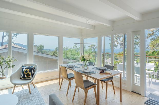 Bright, airy dining area in a contemporary coastal-style home, featuring a timber dining table with upholstered chairs, extensive white-framed glazing, pale timber flooring, and a small adjacent seating area. The windows and glass door open toward a balcony or terrace with leafy outdoor views and water visible in the distance.