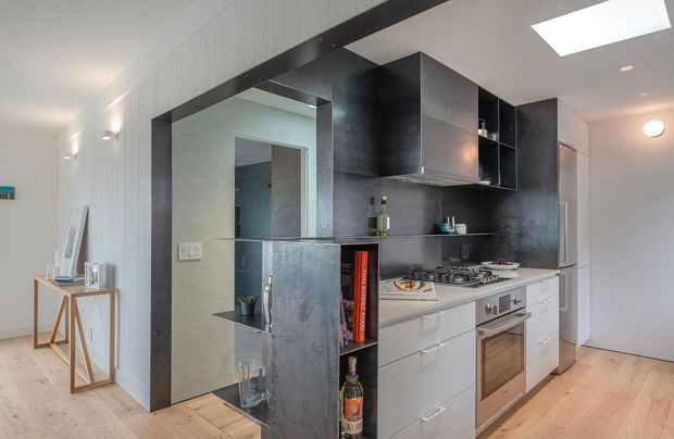 Bright contemporary kitchen with a compact peninsula-style layout, white lower cabinetry, dark charcoal metallic-look upper cabinetry and splashback, integrated stainless-steel appliances, and pale timber flooring. A mirrored or reflective dark-framed opening connects visually to an adjacent hallway or room. White vertical wall panelling, a skylight, and recessed lighting add architectural detail.