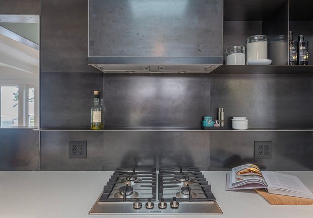 Close-up view of a contemporary kitchen cooking zone with a stainless-steel gas cooktop set into a light benchtop, dark grey metallic wall panels and cabinetry, an integrated rangehood, open shelving with jars and condiments, and a cookbook resting on a wooden board. A window and reflected interior are visible at left.