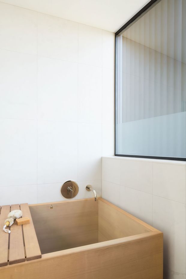 Minimalist bathroom featuring a deep rectangular timber soaking tub with integrated ledge, wall-mounted chrome tapware, large white tiled walls, and a black-framed frosted-glass window. The restrained palette and clean lines create a calm, refined atmosphere.