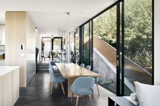Bright contemporary open-plan dining area adjoining a kitchen, with a long light timber dining table and pale blue upholstered chairs. Full-height black-framed glazing runs along one side, overlooking trees and a modern angular exterior structure. Light timber cabinetry, a dark polished floor, pendant lighting, and a partial white sofa create a refined, airy interior.