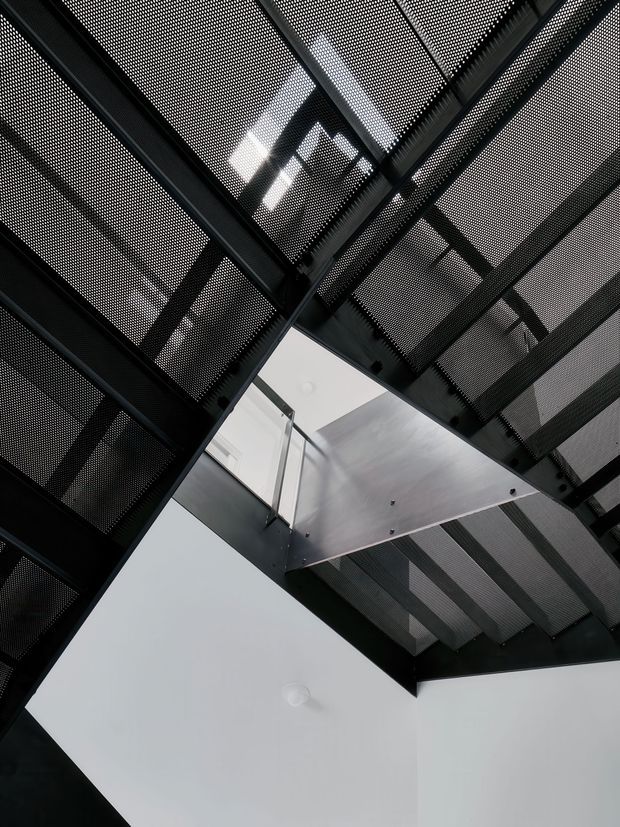 Upward-looking architectural interior view beneath a dramatic black perforated metal ceiling and structural framing. A white geometric ceiling plane and partial glass balustrade create a strong contrast, with visible void-like openings and layered industrial detailing. The image is monochromatic, graphic, and sharply angular.