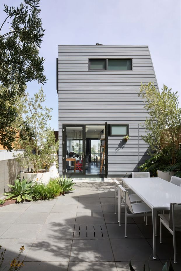 A compact contemporary multi-storey home viewed from a paved courtyard or terrace. The facade is clad in horizontal grey metal or fibre-cement-like panels, with narrow dark-framed windows and large glazed doors opening to the outdoor area. The courtyard includes built-in planters, mature greenery, succulents, and a white outdoor dining table with chairs. Bright daylight and clean geometric lines create a modern urban residential setting.