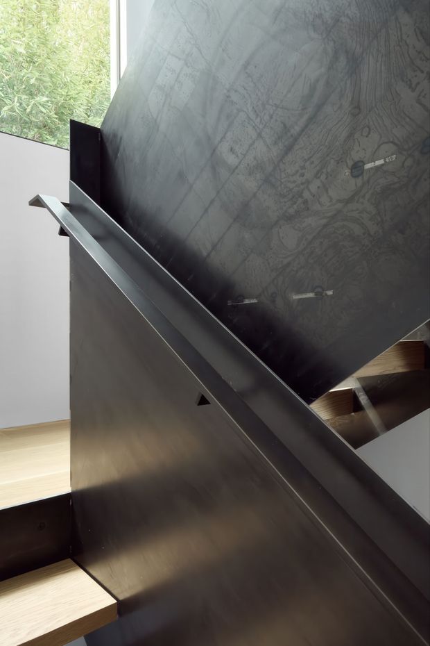 Close-up architectural view of a contemporary staircase with dark metal balustrade and stringer, pale timber treads, and a large dark veined stone or concrete feature wall. A tall window brings in natural light and frames leafy vegetation outside.