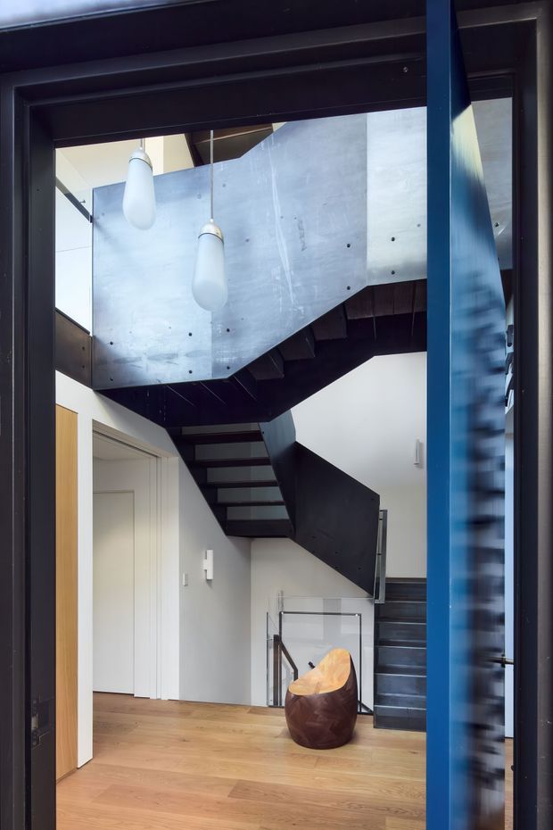 Interior view of a contemporary multi-level stairway and entry hall. A sculptural dark timber or metal staircase wraps around a central void, with a pale concrete or cement-board balustrade, glass railing, white walls, timber flooring, and two suspended pendant lights. A vivid blue framed doorway or structural element appears at the right edge, while a sculptural chair sits below the stairs.