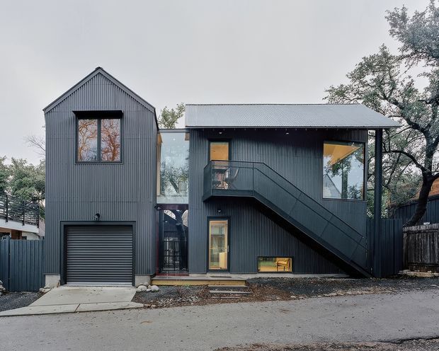 Exterior architectural photograph of a compact multi-level residence with dark grey vertically ribbed metal cladding, a steep gabled volume on the left, and a lower flat-roofed connecting volume on the right. Large glazed openings, a prominent external metal stair, enclosed balcony or circulation space, and an integrated single garage face a paved street. Mature trees and neighbouring structures surround the building.