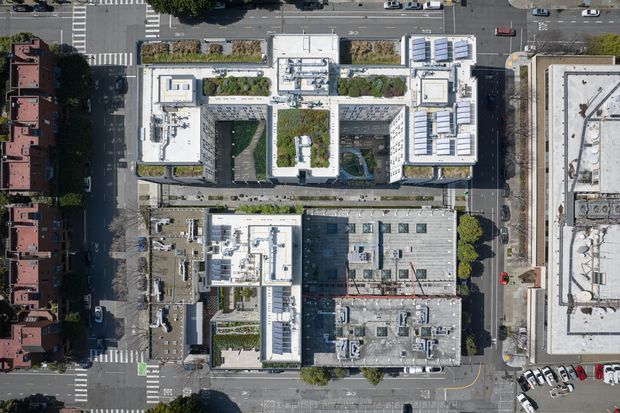 Aerial top-down view of a dense urban block showing several flat-roofed commercial or institutional buildings arranged around landscaped internal courtyards, with rooftop mechanical equipment, solar panels, streets, intersections, and adjacent buildings visible.