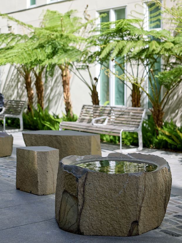 A landscaped courtyard or outdoor terrace beside a light-coloured modern building, featuring a rough-hewn stone water feature or low table in the foreground, matching stone seating blocks, tiled paving, metal slatted benches, and lush fern-like planting. Tall windows and a pale rendered facade form the background. The setting feels calm, organic, and contemporary.
