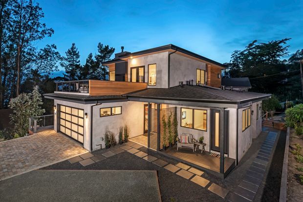 Twilight exterior view of a contemporary multi-level home on a landscaped, sloping site. The house has rendered walls, warm timber cladding, dark low-pitched roof planes, large glazed openings, a covered ground-floor terrace, and an integrated garage with a glazed panel door. Paved paths and outdoor seating surround the building, with mature trees in the background.