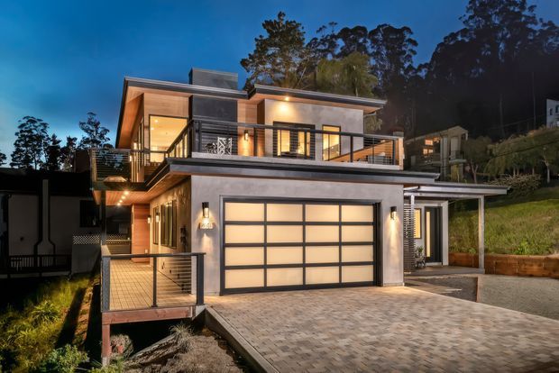 Twilight exterior view of a contemporary multi-level home on a sloping site. The facade combines rendered grey walls, warm timber soffits and cladding, dark-framed glazing, a large frosted-panel garage door, and metal balustrades. A paved driveway, balconies, exterior stairs, and illuminated interior spaces are visible, with trees and neighbouring hillside homes in the background.