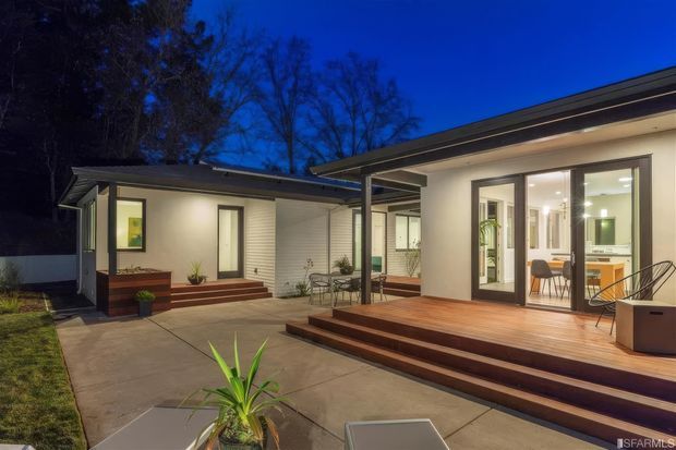 A twilight view of a modern single-storey home arranged around a paved courtyard and timber deck. The house has low-pitched rooflines, light rendered walls, horizontal cladding, extensive glazed doors and windows, and warm interior lighting. Timber steps and decking connect the courtyard to an open-plan interior, with outdoor dining furniture and restrained planting visible.