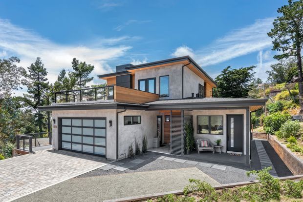 A contemporary multi-level residential home on a sloped, landscaped site. The exterior combines smooth light-grey rendered walls, dark metal roof edging and window frames, warm timber cladding accents, and extensive glazing. A large glazed garage door, upper terrace with glass balustrade, covered entry, paved driveway, and stepped retaining walls are visible.
