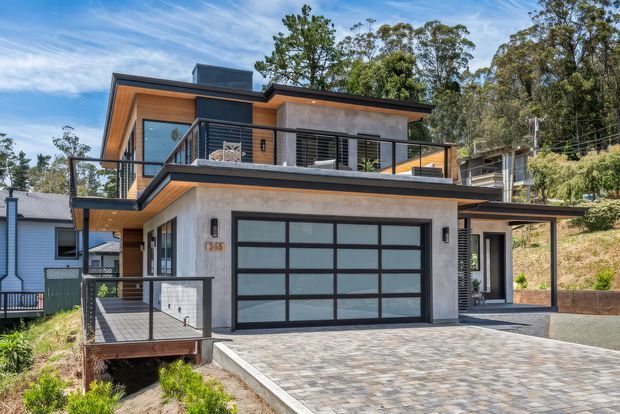 Exterior view of a modern multi-level residence on a sloped, wooded site. The house features a flat roof, projecting upper-storey volumes, concrete-rendered walls, warm timber soffits and cladding, extensive dark-framed glazing, a large glazed sectional garage door, upper-level balconies with black metal railings, and a paved driveway.