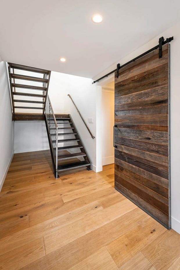 Bright modern-industrial hallway and stairway with wide-plank warm timber flooring, a black metal open-riser staircase and balustrade, white walls, recessed ceiling lights, and a large rustic timber barn-style sliding door on black hardware.