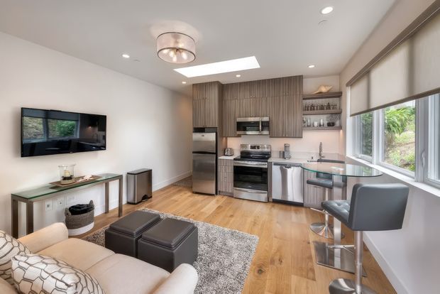 Open-plan living room and kitchen with warm timber flooring, grey vertically grooved cabinetry, stainless-steel appliances, a glass breakfast bar with stools, and large windows fitted with roller blinds. A light sofa, ottomans, textured rug, wall-mounted television, and contemporary pendant light create a bright, compact, modern interior.
