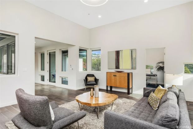 Bright contemporary living room with high ceilings, white walls, large windows and glazed doors, dark timber flooring, and a grey seating arrangement around a round timber coffee table. A textured rug, black occasional chair, timber sideboard, abstract artwork, and indoor plant complete the refined neutral interior.