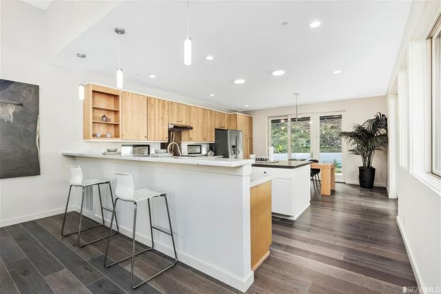 Bright contemporary open-plan kitchen with a large white island and integrated breakfast bar, three white bar stools, light timber cabinetry, dark wood-look flooring, and stainless-steel appliances. The space opens toward a dining area and glazed doors overlooking greenery, with abundant recessed lighting and a potted plant.