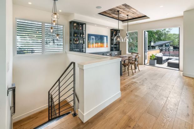 Bright, open-plan residential interior viewed from a stair landing, with warm timber flooring, white walls and cabinetry, a compact bar or kitchen area, dining table, pendant lights, and large glazed doors opening to an outdoor deck. A black metal stair balustrade, built-in bottle storage, and a timber ceiling feature add visual interest.