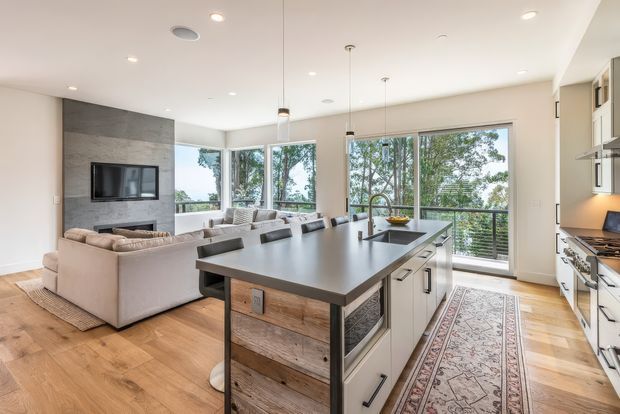 Bright open-plan kitchen and living area with a large central island, white cabinetry, wood flooring, integrated seating, a sectional sofa, and a television set into a dark stone feature wall. Floor-to-ceiling glazing opens to leafy outdoor views and fills the space with natural light.