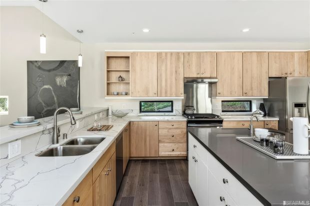 Bright contemporary kitchen with extensive light timber cabinetry, white veined stone benchtops, a large dark island, stainless-steel appliances, pendant lighting, and dark timber flooring. The layout includes a sink run along the left wall and a cooking zone at the rear.