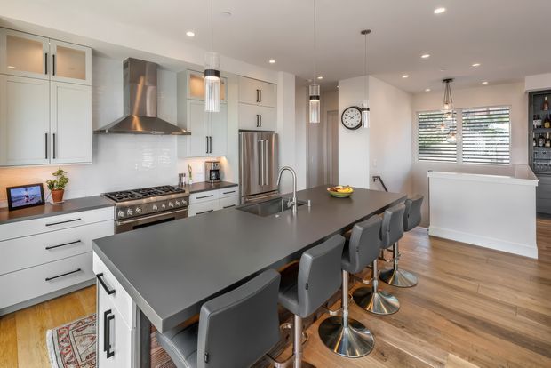 Bright contemporary kitchen with extensive white cabinetry, stainless-steel appliances, a large dark island with integrated sink and seating for several people, pendant lighting, and warm timber flooring. A partial dining or living area and a built-in display/bar cabinet are visible in the background.