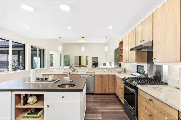 Bright, spacious contemporary kitchen with a large central island, white cabinetry, warm timber wall and base cabinets, dark wood-look flooring, and light stone benchtops. The room features extensive windows, recessed ceiling lighting, stainless-steel appliances, and an open, airy layout.