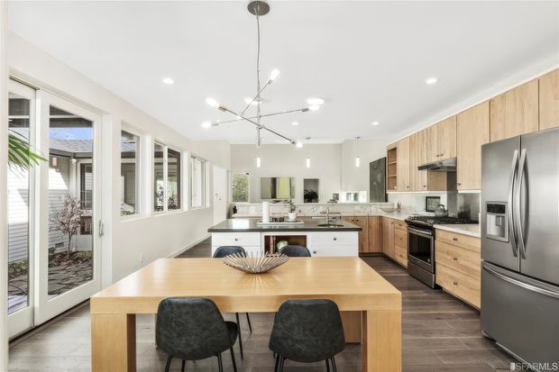 Bright, spacious open-plan kitchen and dining area with extensive natural light, white walls, light timber cabinetry, a central island with seating, stainless-steel appliances, and a timber dining table. Large windows and glazed doors open toward a small courtyard, while contemporary pendant and recessed lighting complete the space.