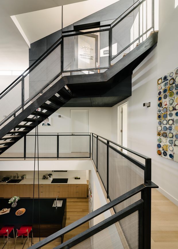 A bright, multi-level interior viewed from an upper landing, featuring a dramatic black open-riser stairway and continuous black metal mesh balustrades. The stairs and landing overlook a contemporary kitchen with light timber cabinetry, a dark island bench, and red stools. White walls, expansive glazing, pale timber flooring, and a colourful circular artwork create a clean, graphic interior composition.