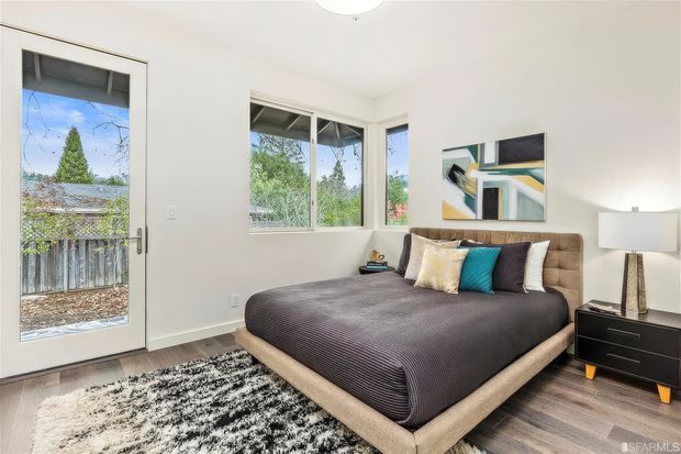 Bright contemporary bedroom with a large upholstered bed, layered neutral and teal bedding, black bedside cabinetry, an abstract wall artwork, and a modern floor lamp. White walls, timber-toned flooring, and expansive corner glazing create an airy connection to the leafy exterior balcony or garden.