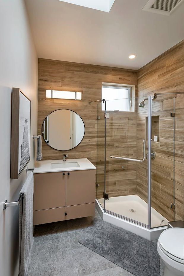 A compact, bright modern bathroom with a wood-look tiled feature wall and matching shower surround. The room includes a floating taupe vanity with white benchtop and integrated basin, round mirror, glass-enclosed accessible shower, wall-mounted fixtures, skylight, recessed lighting, and grey floor tiles.
