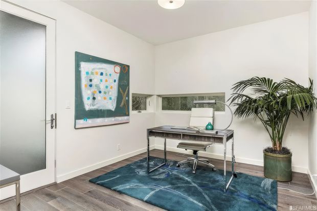 Bright, compact home office with a minimalist metal-and-glass desk, office chair, blue patterned area rug, framed artwork, indoor palm, frosted-glass door, and narrow horizontal windows. The room has white walls and ceiling with dark timber flooring.