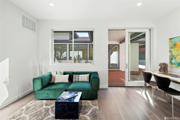 Bright contemporary living room with a deep green sofa, shag rug, dark blue cube coffee table, and a compact dining or work area with black chairs. White walls and ceiling, pale timber flooring, large internal glazing, and a glazed doorway create an open, light-filled interior. A hallway and timber-floored adjoining space are visible beyond the doorway.