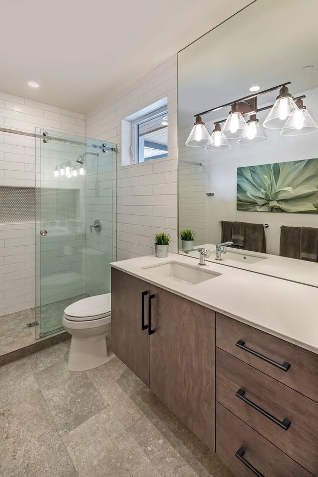 A bright, contemporary bathroom with a glass-enclosed walk-in shower, white subway-tiled walls, a tiled shower niche, toilet, and a broad wood-look vanity with white countertop and integrated sink. A large mirror reflects the dark metal multi-light vanity fixture, while muted botanical artwork and small potted plants add a natural accent.