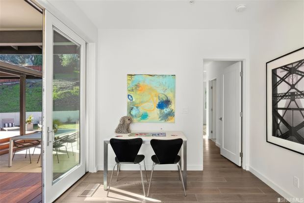 Bright contemporary dining area with a compact white table and two black modern chairs, opening through full-height glazed doors to an outdoor patio. White walls, dark timber flooring, abstract artwork, and a hallway with white panel doors create a clean, light-filled interior.