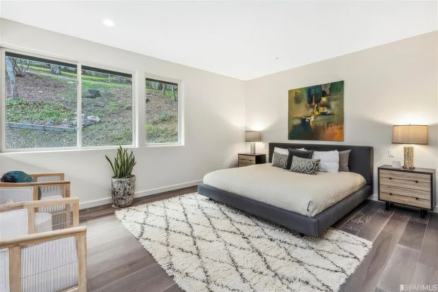 Bright, contemporary bedroom with a large upholstered platform bed, layered neutral bedding, matching bedside tables and lamps, and a large textured geometric rug over dark timber flooring. Floor-to-ceiling-height windows overlook a landscaped slope, while two woven wood-framed lounge chairs and a potted plant add warmth. The room has white walls, a simple recessed ceiling light, and a colourful abstract artwork above the bed.