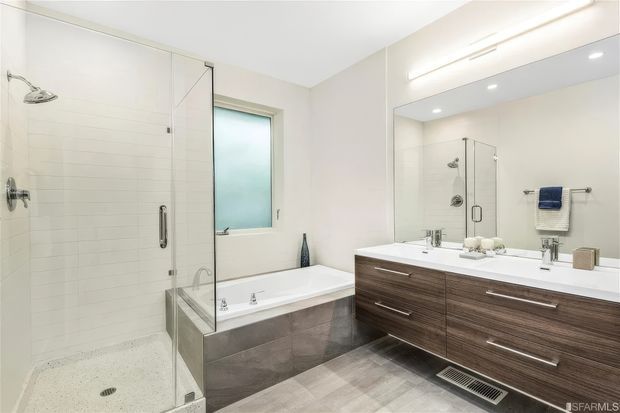 Bright contemporary bathroom with a large frameless glass shower, freestanding-style bathtub set into a tiled surround, and a wide double vanity with dark wood-grain cabinetry and a white benchtop. The room features pale tiled walls, grey floor tiles, chrome fixtures, recessed ceiling lights, and a large mirror.