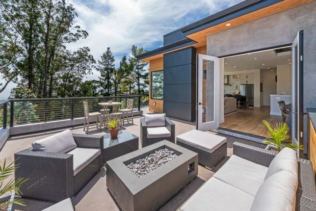 Modern outdoor terrace with a large built-in rectangular fire table, light upholstered outdoor seating, woven dark-grey furniture, and a dining setting. The terrace has horizontal metal cable-style balustrading, timber soffit accents, dark panel cladding, and opens directly into a contemporary kitchen and living area. Mature trees and a sloping natural setting are visible beyond the railing.