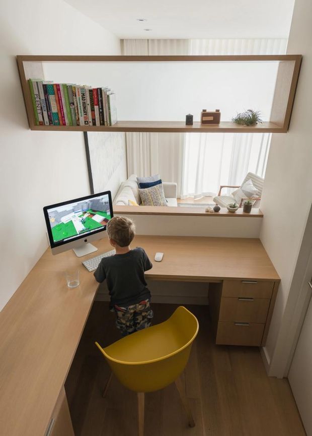 A compact built-in home office viewed from above, with warm timber desk surfaces wrapping around the room, integrated drawers, a computer workstation, and a mustard-yellow chair. A large timber-framed interior window with a narrow display shelf looks into a bright sitting area, with books, small decor, and plants arranged along the shelf. Sheer curtains and recessed ceiling lights create a bright, clean workspace.