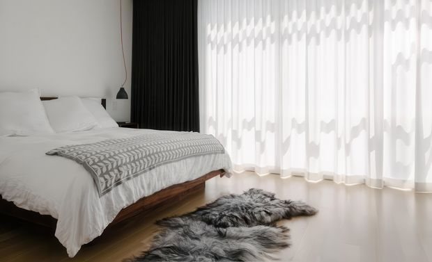 A bright, minimalist bedroom with a neatly made bed, white bedding, a grey patterned throw, and a dark timber bed base. Full-height sheer curtains diffuse abundant daylight beside dark blackout drapes, while layered grey faux-fur rugs add texture on the pale floor. A small black pendant light hangs near the bedside.