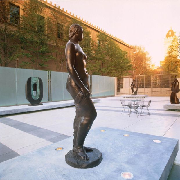 A landscaped urban courtyard or public plaza featuring a prominent bronze figurative sculpture on a pedestal, additional sculptures, pale stone paving, outdoor café-style tables and chairs, and a long brick building with vertical greenery and decorative roofline details in the background. Warm late-afternoon light creates a refined outdoor gallery atmosphere.