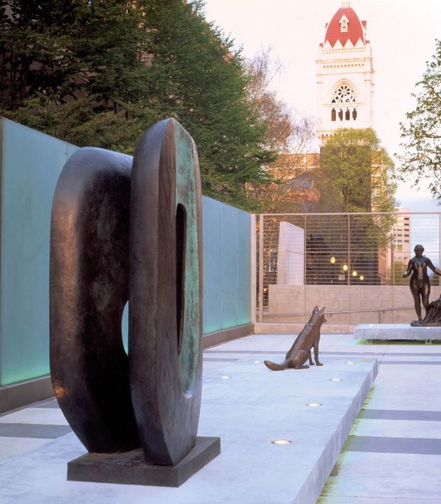 A landscaped urban courtyard or sculpture garden featuring a large dark bronze abstract sculpture in the foreground, smaller figurative sculptures along a pale paved pathway, turquoise privacy walls, and integrated circular ground lights. A distinctive historic tower rises behind the courtyard, partially framed by mature trees.