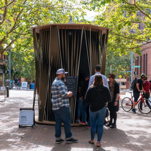 A temporary or compact architectural pavilion stands in a leafy urban pedestrian plaza. The structure has a rounded, slightly tapered form with a dark interior and a sculptural facade made from closely spaced vertical timber-like slats, topped by a shallow curved roof. Several pedestrians gather around and enter the pavilion, while bicycles and brick civic or commercial buildings are visible in the surrounding streetscape.