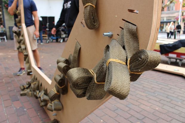 Close-up of a temporary outdoor installation or exhibit featuring a large curved wood-toned panel with numerous rolled and bundled woven or braided textile elements attached to its surface. People and additional display structures are visible in the brick-paved public setting behind it.