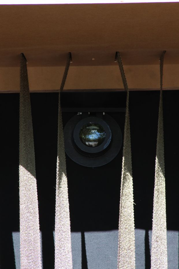 Close-up architectural detail showing a dark facade or wall with a circular porthole-style window, partially screened by several hanging woven or mesh vertical elements. A warm-toned horizontal soffit or overhang spans the top, with strong sunlight creating high-contrast shadows across the facade.